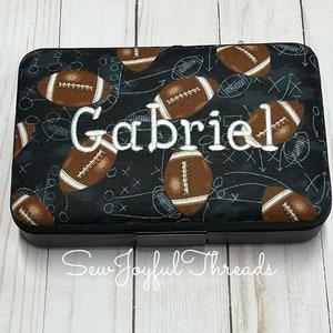 May include: A black zippered pouch with a football pattern and the name "Gabriel" embroidered in white. The pouch is on a white wooden surface.