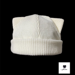 May include: Cream-colored knit beanie with cat ears. The hat features a ribbed design on the brim and a smooth texture on the crown. The cat ears are upright and add a playful touch. The background is black.