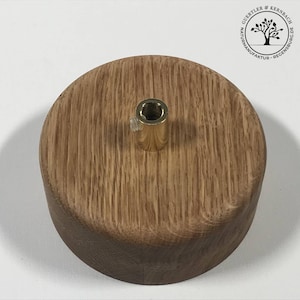 Puede incluir: Una base redonda de madera con un inserto roscado de latón en el centro. La base está hecha de madera marrón claro con un acabado natural.