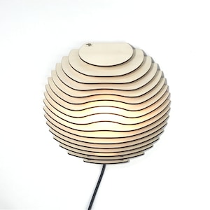 Könnte beinhalten: Eine Wandlampe aus Holz mit einem geschichteten, abgerundeten Design. Die Lampe ist beleuchtet und strahlt ein warmes Licht aus. Die Lampe hat ein schwarzes Kabel.