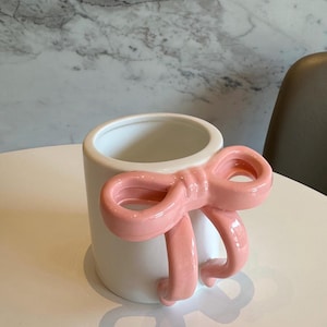 May include: A white ceramic mug with a large, pink bow-shaped handle. The bow is a solid, glossy pink, and the mug has a simple, cylindrical shape. The mug sits on a white surface.
