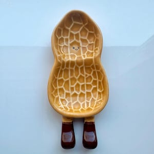 May include: A whimsical peanut-shaped spoon rest with a smiling face. The spoon rest is tan with a textured surface and dark brown legs. A fun and functional kitchen accessory.