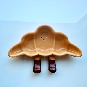 May include: A tan ceramic cloud-shaped dish with two dark brown boot-shaped legs. The dish has a smiling face and is designed for serving food or displaying small items. The dish is on a light blue surface.