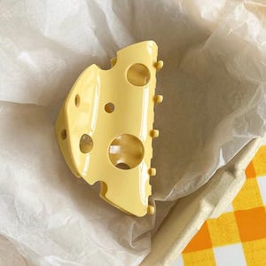 May include: A pale yellow hair claw clip shaped like a wedge of Swiss cheese, featuring a smooth, glossy finish and several circular cutouts of varying sizes across its surface, with small teeth along one edge for securing hair in place.
