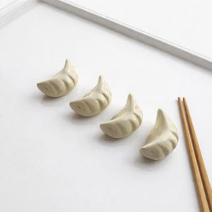 May include: Four off-white ceramic dumpling chopstick rests arranged in a row on a white surface. Two wooden chopsticks are placed to the right of the dumpling rests. The dumpling rests are a light beige colour.