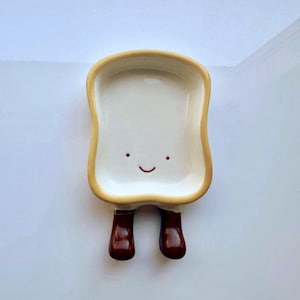 May include: A novelty ceramic dish in the shape of a slice of bread, featuring a smiling face and brown legs. The dish has a white interior and a yellow-brown crust. Ideal for holding small items, it's a fun decorative piece.