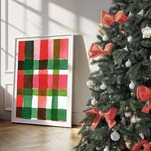 May include: A framed abstract art print with a plaid pattern in red, green, and brown hues. A decorated Christmas tree with red bows and ornaments stands nearby. The artwork leans against a wall.