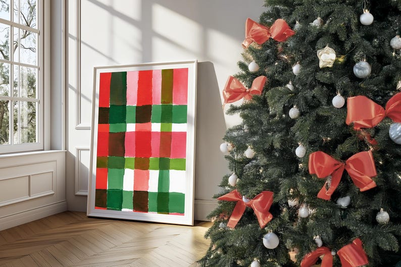 May include: A framed abstract art print with a plaid pattern in red, green, and brown hues. A decorated Christmas tree with red bows and ornaments stands nearby. The artwork leans against a wall.