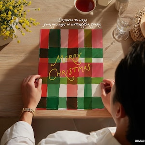 May include: A Christmas gift wrap with a plaid design in red, green, and pink. The words "MERRY CHRISTMAS" are written in yellow script. The image also includes text that says "DESIGNED TO WRAP your holidays in watercolor charm."