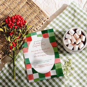 May include: A Christmas party invitation with a watercolour design in red, green, and white. The invitation is next to a cup of hot chocolate topped with marshmallows and a sprig of red berries. A woven basket is in the background.