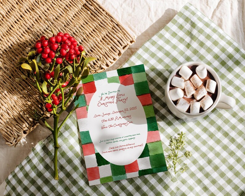 May include: A Christmas party invitation with a watercolour design in red, green, and white. The invitation is next to a cup of hot chocolate topped with marshmallows and a sprig of red berries. A woven basket is in the background.