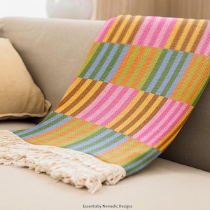 May include: A colorful woven throw blanket with a striped pattern in shades of pink, yellow, green, blue, and brown. The blanket has a fringed edge and is draped over a beige sofa. The text "Essentially Nomadic Designs" is visible.