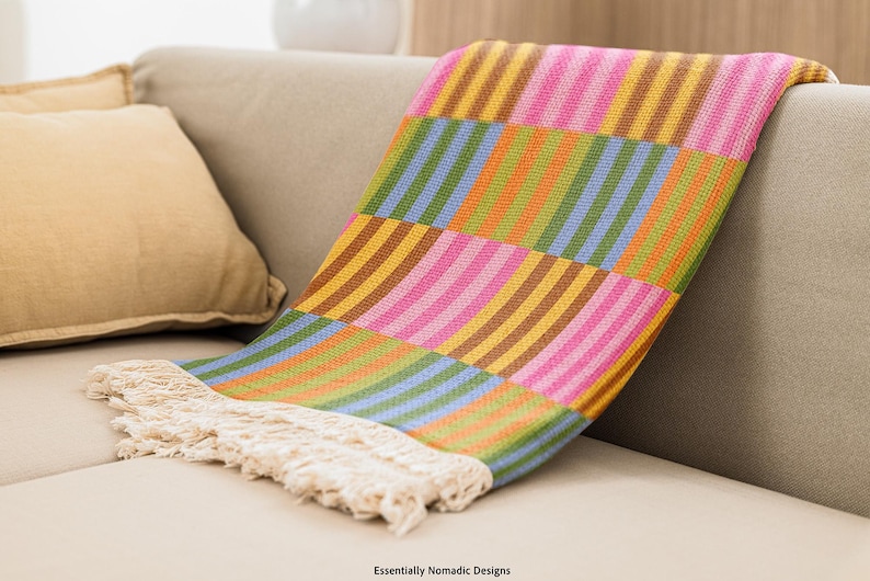 May include: A colorful woven throw blanket with a striped pattern in shades of pink, yellow, green, blue, and brown. The blanket has a fringed edge and is draped over a beige sofa. The text "Essentially Nomadic Designs" is visible.