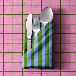 May include: A stainless steel knife, fork, and spoon are neatly arranged in a folded napkin with blue and green stripes. The napkin rests on a pink checkered tablecloth. The text "essentially nomadic designs" is visible.