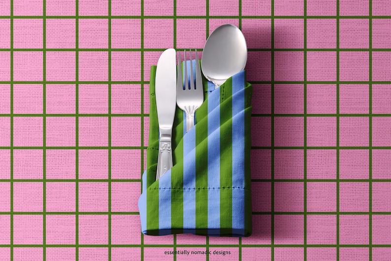 May include: A stainless steel knife, fork, and spoon are neatly arranged in a folded napkin with blue and green stripes. The napkin rests on a pink checkered tablecloth. The text "essentially nomadic designs" is visible.