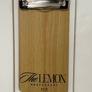 May include: A wooden clipboard with a metal clip and the text "The Lemon Restaurant EstD 2023" printed on it.