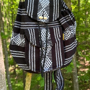 May include: Black and white woven backpack with a flap closure and drawstring. The backpack features a geometric pattern and wooden toggle closures. The bag is hanging on a tree branch in a natural outdoor setting.