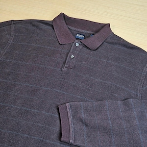 May include: A dark brown long-sleeve polo shirt featuring a subtle grid pattern in a lighter shade. The shirt has a classic collar, a two-button placket with dark buttons, and ribbed cuffs. An Arrow brand label is visible inside the collar.