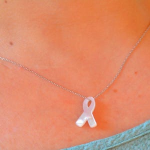 Può includere: Una collana d'argento con un ciondolo a forma di nastro bianco. Il nastro è un simbolo di consapevolezza. La collana è mostrata su uno sfondo color carne e un top blu.
