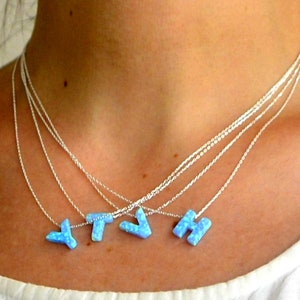Puede incluir: Un conjunto de collares superpuestos con cuatro cadenas finas de plata, cada una con un colgante de letra azul claro estilo ópalo. Los colgantes muestran las letras Y, T, V y H, con una textura moteada e iridiscente en tono azul.