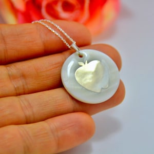 May include: A silver necklace featuring a round, white pendant with a silver apple design. The pendant is held by a hand, showcasing the necklace's details. The chain is delicate and silver.