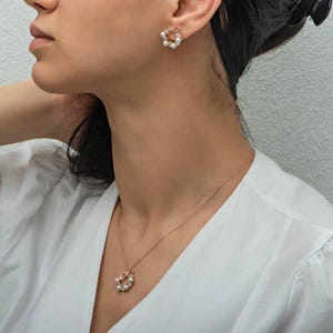 Peut inclure: Un ensemble collier et boucles d'oreilles à clous en or rose, composé d'un motif couronne circulaire orné de petites perles blanches et de cristaux transparents, avec un petit nœud sur le dessus, porté sur une chemise blanche.