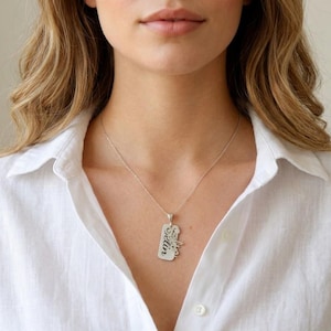 Puede incluir: Un collar con colgante de plata con una etiqueta rectangular y un diseño de nombre en cursiva. El collar se lleva sobre una camisa blanca abotonada. El colgante tiene un pequeño elemento decorativo.