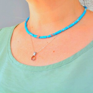 Può includere: Un set di collane a strati composto da un filo corto di perline cilindriche blu brillante con una sottile venatura marrone e una delicata catenina color argento con un ciondolo a forma di pera di colore ambra traslucido.