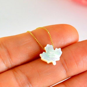 May include: A necklace featuring a white, iridescent maple leaf pendant suspended from a thin gold-colored chain. The pendant displays a smooth, pearlescent surface with subtle light-reflecting qualities, shaped into the distinct form of a maple leaf.