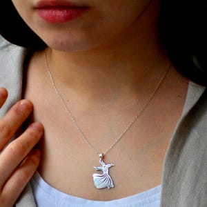 Pode incluir: Um colar com pingente de prata com um design de dervixe rodopiante. O pingente é suspenso por uma corrente delicada. O colar é usado sobre uma parte superior de cor clara.