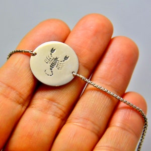 May include: A silver bracelet with a circular pendant engraved with a scorpion symbol. The pendant is attached to a fine chain. The bracelet is displayed against a plain backdrop.