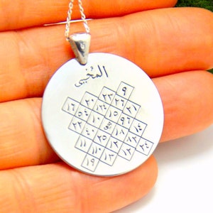 May include: A silver pendant necklace with a round charm. The charm features Arabic script and a grid of numbers. The pendant hangs from a delicate silver chain. The background is a person's hand.