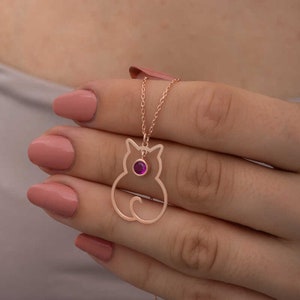 Puede incluir: Collar con colgante de gato en oro rosa con una pequeña gema redonda de color magenta. El colgante de gato es un contorno simple con una cola rizada. El collar está en una cadena delicada.
