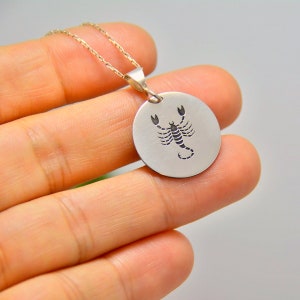 May include: A silver pendant necklace with a circular charm displaying a detailed scorpion design. The scorpion is black and centrally placed. The chain is delicate silver. The pendant is held by a person's hand.