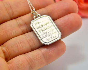 May include: A rectangular silver pendant necklace with beveled edges and a polished finish. The center of the pendant features intricate, engraved Arabic calligraphy in a dark, contrasting tone, suspended from a delicate silver chain.