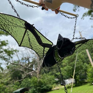 May include: A stained glass bat suncatcher featuring textured light green wings and a solid black body. The bat is constructed with dark metal solder lines and is suspended by a thin black metal chain attached to a wooden dowel for display.