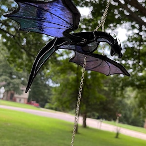 May include: A black stained glass dragon with iridescent blue and purple wings hangs from a silver chain. The dragon has a detailed design and a crystal pendant at the bottom. The background is a blurred outdoor scene.