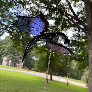 Może przedstawiać: Czarny łapacz słońca w kształcie smoka z iryzującymi niebieskimi i fioletowymi skrzydłami, zawieszony na srebrnym łańcuszku. Smok zawieszony jest na tle rozmytego zielonego tła, z przezroczystym kryształowym wisiorkiem poniżej.