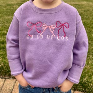 May include: A purple sweatshirt with three pink and red bows embroidered on the front. The text "CHILD OF GOD" is embroidered below the bows.