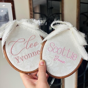 May include: Two circular wooden embroidery hoops containing white fabric with a subtle floral pattern. The left hoop is embroidered with the names Cleo and Yvonne in pink thread. The right hoop features the names Scottie and Jane in pink.