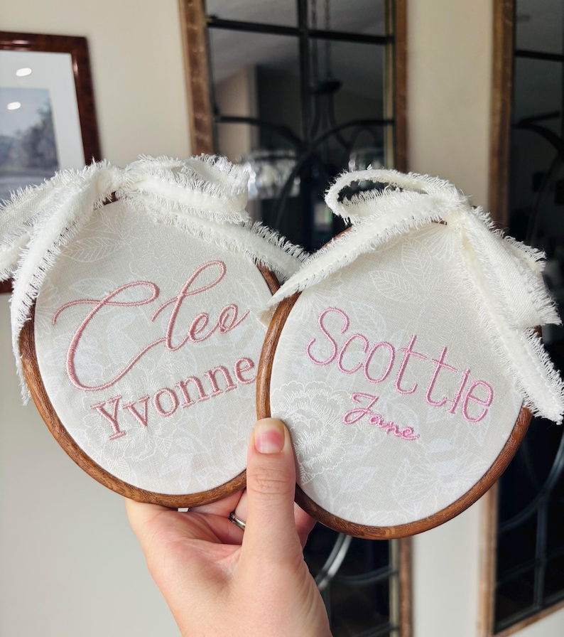 May include: Two circular wooden embroidery hoops containing white fabric with a subtle floral pattern. The left hoop is embroidered with the names Cleo and Yvonne in pink thread. The right hoop features the names Scottie and Jane in pink.