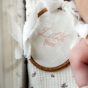 May include: A circular wooden embroidery hoop containing white fabric with a subtle floral pattern. The name Lilah and the word June are embroidered in pink thread. A white frayed ribbon is tied to the top of the wooden frame.