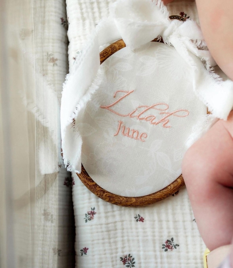 May include: A circular wooden embroidery hoop containing white fabric with a subtle floral pattern. The name Lilah and the word June are embroidered in pink thread. A white frayed ribbon is tied to the top of the wooden frame.
