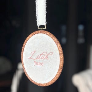 May include: An oval embroidery hoop with a faux wood grain frame containing white fabric with a subtle floral pattern. The name Lilah and the month June are embroidered in pink thread. A white frayed ribbon is attached to the top for hanging.
