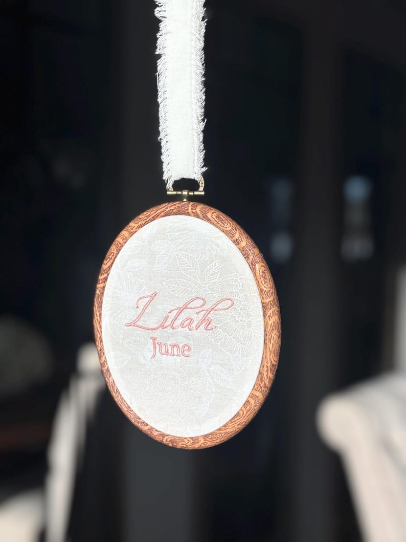 May include: An oval embroidery hoop with a faux wood grain frame containing white fabric with a subtle floral pattern. The name Lilah and the month June are embroidered in pink thread. A white frayed ribbon is attached to the top for hanging.