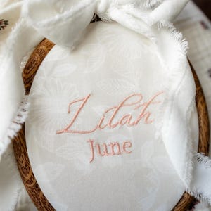 May include: An oval wooden embroidery hoop containing white fabric with a subtle white floral pattern. The name Lilah and the month June are embroidered in pink thread. A white fabric ribbon with frayed edges is tied to the top of the hoop.