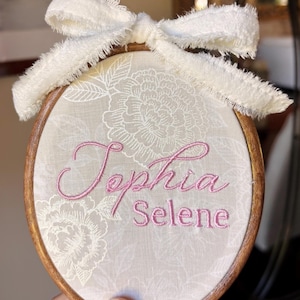 May include: An oval wooden embroidery hoop containing beige fabric with a white floral pattern. The names Tephia and Selene are embroidered in pink thread across the center. A frayed white ribbon is tied in a bow at the top.