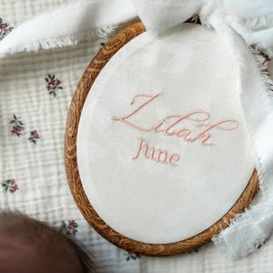May include: An oval wooden embroidery hoop containing white fabric with a subtle floral pattern. The name Lilah and the month June are embroidered in light pink thread. A white frayed ribbon is tied to the top of the wooden frame.