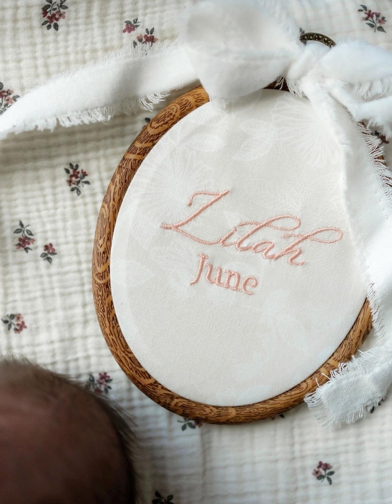 May include: An oval wooden embroidery hoop containing white fabric with a subtle floral pattern. The name Lilah and the month June are embroidered in light pink thread. A white frayed ribbon is tied to the top of the wooden frame.