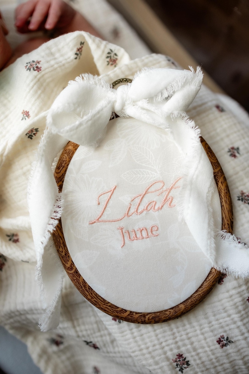 May include: An oval wooden embroidery hoop containing white fabric with a subtle floral pattern. The name Lilah and the month June are embroidered in pink thread. A white frayed ribbon is tied to the top of the hoop.